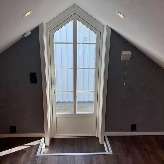 Innvendig visning av spiss loftsdør i hvit karm og mørkt gulv.